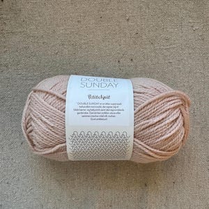 May include: A ball of light pink yarn with a white label that reads "DOUBLE SUNDAY" and "Petiteknit." The label includes text in a foreign language and a decorative pattern. The yarn is likely for knitting or crocheting.