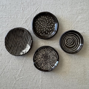 May include: Four small, round, black ceramic dishes with white hand-painted designs. One dish features vertical lines, another has a sunburst pattern, one displays a central flower motif, and the final dish shows a series of concentric circles.