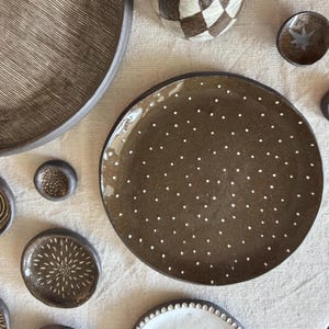 May include: A collection of dark brown ceramic tableware including a large plate with white polka dots, a bowl with fine linear etchings, and several small dishes featuring various white patterns like starbursts and dots on a textured surface.