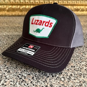 Op de afbeelding: Zwarte truckerpet met een grijze mesh achterkant. De voorkant heeft een patch met het woord "Lizards" in het rood, een groene dinosaurusafbeelding en een groene rand. De pet heeft een gebogen klep en een verstelbare band.