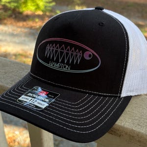 May include: Black and white trucker hat with the word "HAMPTON" in an oval design. The design has a fish-like graphic with a gradient of pink, purple, and blue. The hat has a black brim and crown, and a white mesh back.