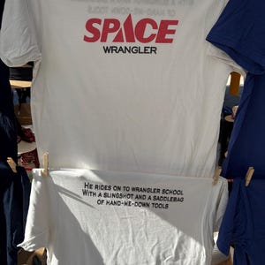 May include: Two white t-shirts are displayed. The top shirt has the word "SPACE" in red and "WRANGLER" in black. The bottom shirt has the text: "HE RIDES ON TO WRANGLER SCHOOL WITH A SLINGSHOT AND A SADDLEBAG OF HAND-ME-DOWN TOOLS."