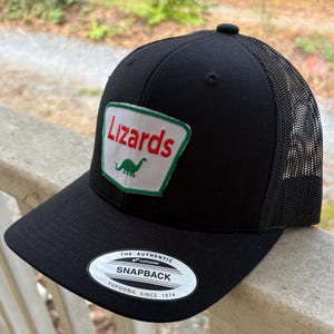 Könnte beinhalten: Schwarze Snapback-Trucker-Mütze mit Mesh-Rückseite. Auf der Vorderseite befindet sich ein Aufnäher mit dem Wort "Lizards" in Rot und einer grünen Dinosaurier-Grafik. Die Mütze hat einen gebogenen Schirm und einen Schnappverschluss.