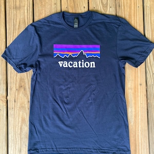Könnte beinhalten: Ein marineblaues T-Shirt mit einer weißen Grafik einer Bergkette und dem Wort "vacation" darunter. Die Grafik ist vom Patagonia-Logo inspiriert.