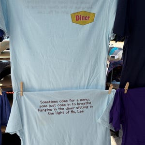 May include: Light blue t-shirts hanging on a clothesline. One shirt has a Diner logo and text. The other shirt has text that reads, "Sometimes come for a menu, some just come in to breathe Hanging in the diner sitting in the light of Ms. Lee."
