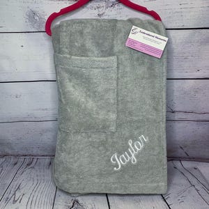May include: A gray bath towel with a pocket, hanging on a pink hanger. The name "Taylor" is embroidered in white script. A small business card with the text "Embroidered Memories" is attached.