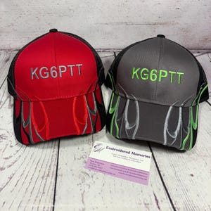 May include: Two trucker hats, one red and one gray, with black mesh backs. Each hat features the embroidered letters "KG6PTT" and flame designs in contrasting colors. The red hat has gray flames, and the gray hat has green flames.