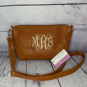 May include: A tan crossbody bag with a textured surface and a gold embroidered monogram featuring the letters MRJ in a script font. The bag includes a matching adjustable shoulder strap with gold-toned metal hardware and a small business card.