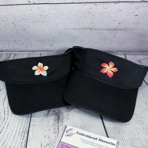 May include: Two black visors featuring embroidered plumeria flower designs. The left visor has a white and yellow flower, while the right visor displays a pink and orange flower. A business card labeled Embroidered Memories is visible below.