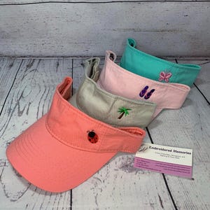 May include: Four colorful visors in coral, tan, pink, and teal. Each visor has an embroidered design: a ladybug, palm tree, flip-flops, and butterfly. The visors are displayed on a light wood-grain surface.