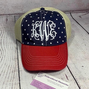 May include: A baseball cap featuring a navy blue front panel with white stars and a white embroidered monogram of the letters KWE. The cap has a red brim with white stitching and a tan mesh back panel. A business card is placed in front of the cap.