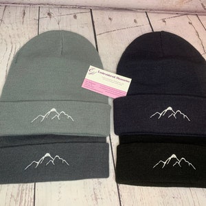May include: Two knit beanies with a white mountain embroidered design. The beanie on the left is gray and the beanie on the right is black.