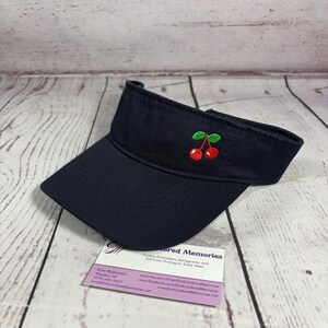 May include: A navy blue visor featuring an embroidered design of two red cherries with green leaves on the front panel. The visor has a curved brim and an adjustable strap. A business card for Embroidered Memories is placed underneath.