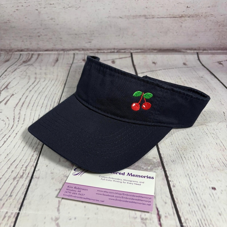 Puede incluir: Una visera azul marino con un dise&ntilde;o bordado de dos cerezas rojas con hojas verdes en el panel frontal. La visera tiene una visera curva y una correa ajustable. Debajo se encuentra una tarjeta de visita de Embroidered Memories.