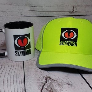 May include: A white ceramic mug with a black handle and interior, and a neon green trucker hat. Both items display a black square with a red and white eye design and the word "SKYWARN" in black text. The hat has a grey brim.