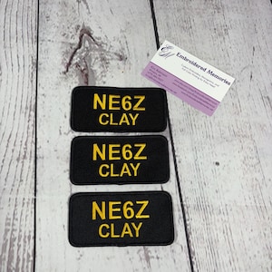 May include: Three rectangular black fabric patches with embroidered yellow text reading NE6Z CLAY. A white business card with the text Embroidered Memories, Kim Robinson, and contact details is positioned above the patches.