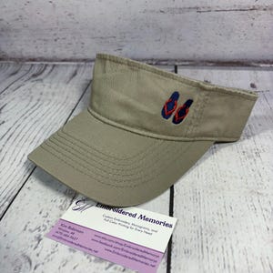 Puede incluir: Una visera de color caqui con un dise&ntilde;o bordado de un par de chanclas azul marino con tiras rojas. La visera tiene un ala estructurada con costuras visibles y un dise&ntilde;o abierto en la parte superior, ofreciendo una opci&oacute;n de accesorio simple.