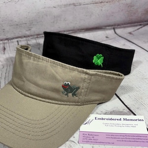 Puede incluir: Una visera color canela con una rana gris bordada de boca roja y una visera negra con un tr&eacute;bol de cuatro hojas verde bordado. Una tarjeta de visita de Embroidered Memories con informaci&oacute;n de contacto se encuentra junto a las dos viseras.