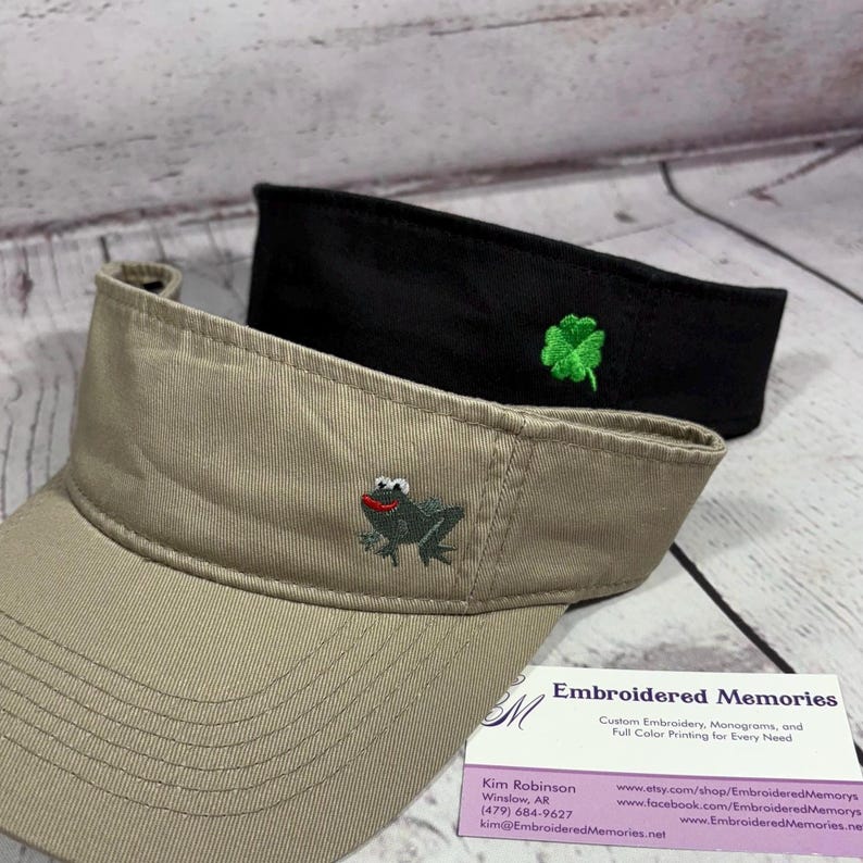 Puede incluir: Una visera color canela con una rana gris bordada de boca roja y una visera negra con un tr&eacute;bol de cuatro hojas verde bordado. Una tarjeta de visita de Embroidered Memories con informaci&oacute;n de contacto se encuentra junto a las dos viseras.