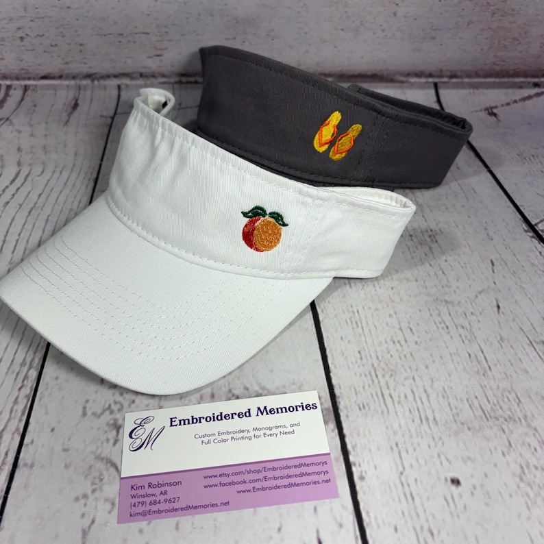 Puede incluir: Una visera blanca con un melocot&oacute;n bordado y una visera gris oscuro con chanclas amarillas bordadas. Una tarjeta de visita de Embroidered Memories con informaci&oacute;n de contacto est&aacute; colocada frente a las dos viseras sobre madera clara.