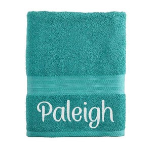 May include: A rectangular teal bath towel with a textured terry cloth surface. The towel features a horizontal band with decorative ribbed detailing and the name Paleigh embroidered in white script lettering across the lower section.