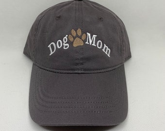 May include: A gray baseball cap with the embroidered text "Dog Mom" in white, arched above a tan paw print. The cap has a curved brim and a button on top.