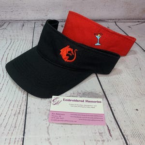 Puede incluir: Una visera negra con un drag&oacute;n rojo bordado y una visera roja con una copa de martini bordada. Se muestra una tarjeta de visita de Embroidered Memories con datos de contacto, el nombre Kim Robinson y la direcci&oacute;n del sitio web.