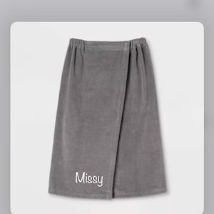 May include: A gray wrap towel with an elastic waistband. The towel is made of a soft material and features the name "Missy" in white script near the bottom. The towel is designed to be worn around the body.