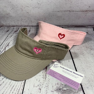 Puede incluir: Dos viseras, una de color verde oliva y otra de color rosa claro, cada una con un peque&ntilde;o dise&ntilde;o de coraz&oacute;n bordado en la parte central delantera. Una tarjeta de visita de Embroidered Memories con datos de contacto est&aacute; junto a las viseras.
