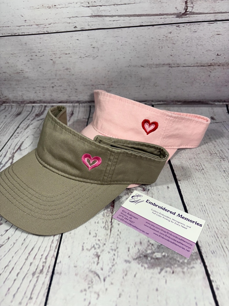 Puede incluir: Dos viseras, una de color verde oliva y otra de color rosa claro, cada una con un peque&ntilde;o dise&ntilde;o de coraz&oacute;n bordado en la parte central delantera. Una tarjeta de visita de Embroidered Memories con datos de contacto est&aacute; junto a las viseras.