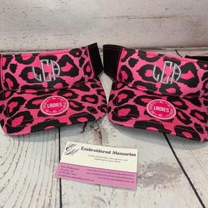 May include: Two pink and black leopard print visors with the initials "LEA" embroidered on the front. The visors have a black band and a white circle with the word "LADIES" in black lettering.