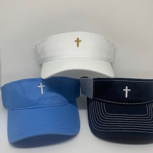 May include: Three visors in white, light blue, and navy blue, each featuring a cross emblem. The white visor has a gold cross, while the others have white crosses. The visors are made of a durable fabric.