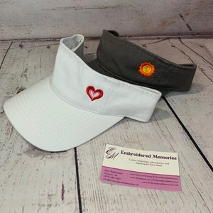 Puede incluir: Una visera blanca con un coraz&oacute;n bordado en rojo y rosa y una visera gris oscuro con un sol bordado en amarillo y naranja. Una tarjeta de visita de Embroidered Memories con datos de contacto e informaci&oacute;n del sitio web est&aacute; en primer plano.