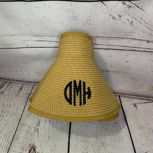 May include: A tan straw sun visor featuring a wide brim and a structured crown. The front of the visor is embroidered with a black circular monogram featuring the letters DMH in a stylized, bold font with vertical line detailing.