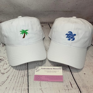 May include: Two white baseball caps featuring embroidered designs. The left cap displays a green and brown palm tree, while the right cap features a blue sea turtle. A business card for Embroidered Memories is positioned between the two hats.