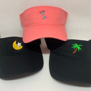 May include: Three visors in the image. One is coral with a martini embroidery, another is black with a moon and star, and the third is black with a palm tree. All visors have a curved brim and are made of a sturdy fabric.