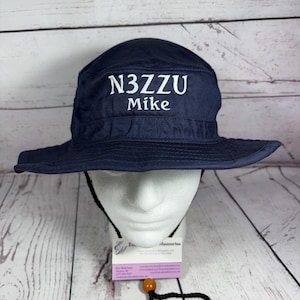 May include: A navy blue bucket hat featuring white embroidered text that reads N3ZZU and Mike on the front. The hat includes a black chin strap with an adjustable orange bead toggle, designed with a wide brim and a structured crown.