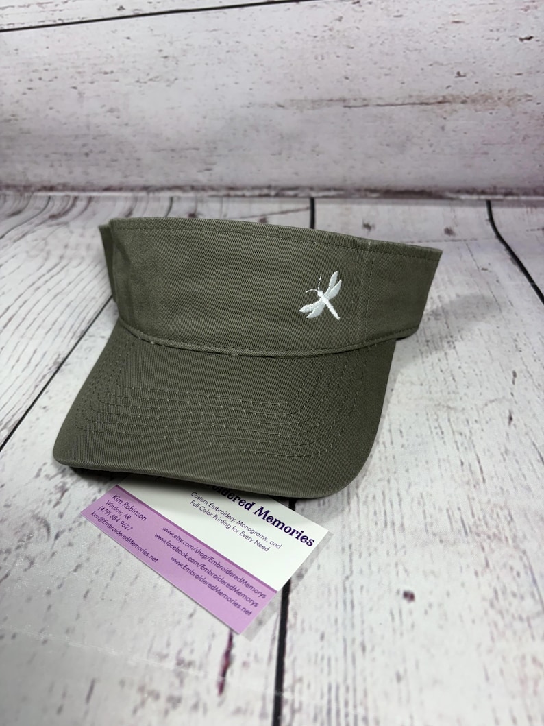 Puede incluir: Una visera de color verde oliva con una lib&eacute;lula blanca bordada en el panel frontal. Debajo se encuentra una peque&ntilde;a tarjeta de visita blanca con el texto Embroidered Memories, Kim Robinson, Winslow, AR, (479) 684-9627 y detalles web.