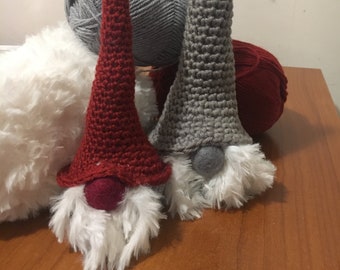 Pode incluir: Dois gnomos de crochê feitos à mão com chapéus longos e pontudos. Um gnomo tem um chapéu vermelho e o outro cinza. Ambos têm barbas brancas e fofas e narizes pequenos e redondos. Bolas de lã cinza, branca e vermelha no fundo.