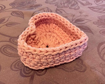 May include: A heart-shaped crochet basket crafted from thick, peach-colored cotton yarn. The container features a textured, woven stitch pattern with raised edges and a hollow center, forming a small, open-top bowl structure.