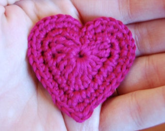 May include: A handmade, magenta-colored crochet heart. The heart is held in an open hand, showcasing the detailed stitch work. The heart's shape is clearly defined, with a rounded top and a pointed bottom, suggesting a Valentine's Day or craft theme.