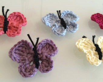 May include: Five crocheted butterflies in different colors: pink, purple, blue, cream, and red. The butterflies have black yarn for the antennae and body.