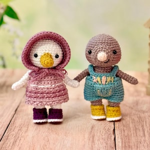 May include: Two crocheted stuffed toys, a white bird wearing a pink dress and a brown mole wearing blue overalls. The toys are standing on a wooden surface.