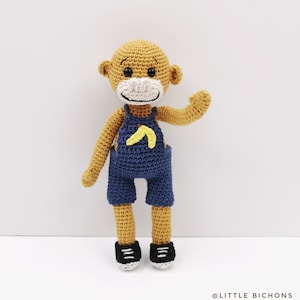 May include: A crocheted monkey doll wearing blue overalls with a yellow banana design. The monkey has a white face with black eyes and a black mouth.