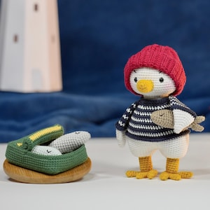Puede incluir: Muñeca de gaviota de ganchillo hecha a mano con gorro rojo y suéter a rayas azul marino y blanco, sosteniendo un pez. Una cesta de ganchillo verde con peces se encuentra sobre una base de madera, con un faro al fondo.