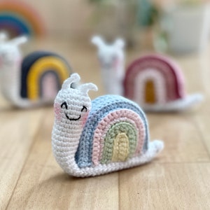 May include: Three crocheted snail toys with rainbow shells. The snails are white with black eyes and smiling faces. The rainbow shells are different colors: black, red, and blue.