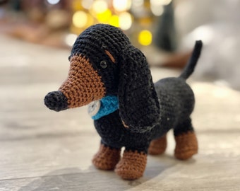 May include: A crocheted dachshund dog with a black body, brown snout, and paws, and a blue collar. The handmade stuffed animal is standing on a wooden surface. The dog has a long body and short legs.