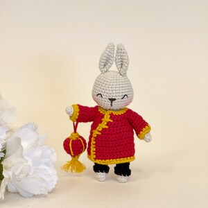 May include: A white crocheted bunny wearing a red and yellow Chinese-style robe and holding a red and yellow lantern.