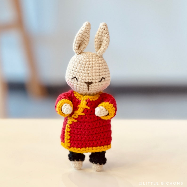 May include: A crocheted rabbit wearing a red and yellow Chinese-style outfit. The rabbit has a white face with black eyes and a brown nose.