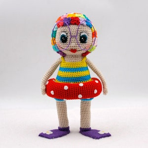 Peut inclure: Une poupée en crochet portant un maillot de bain coloré, une bouée de sauvetage rouge à pois blancs et des lunettes violettes. La poupée a une tête en forme de fleur colorée.